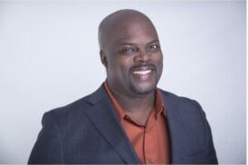 A man with a shaved head, goatee, and mustache smiles at the camera in this actor headshot. He wears a dark blazer over an orange shirt, standing against a plain light background—perfect for actor headshots Calgary style. - Jodi O Photography