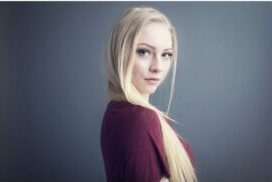 A young woman with long blonde hair, wearing a maroon shirt, stands against a plain gray background, looking over her shoulder toward the camera with a neutral expression—perfect for professional actor headshots Calgary style. - Jodi O Photography