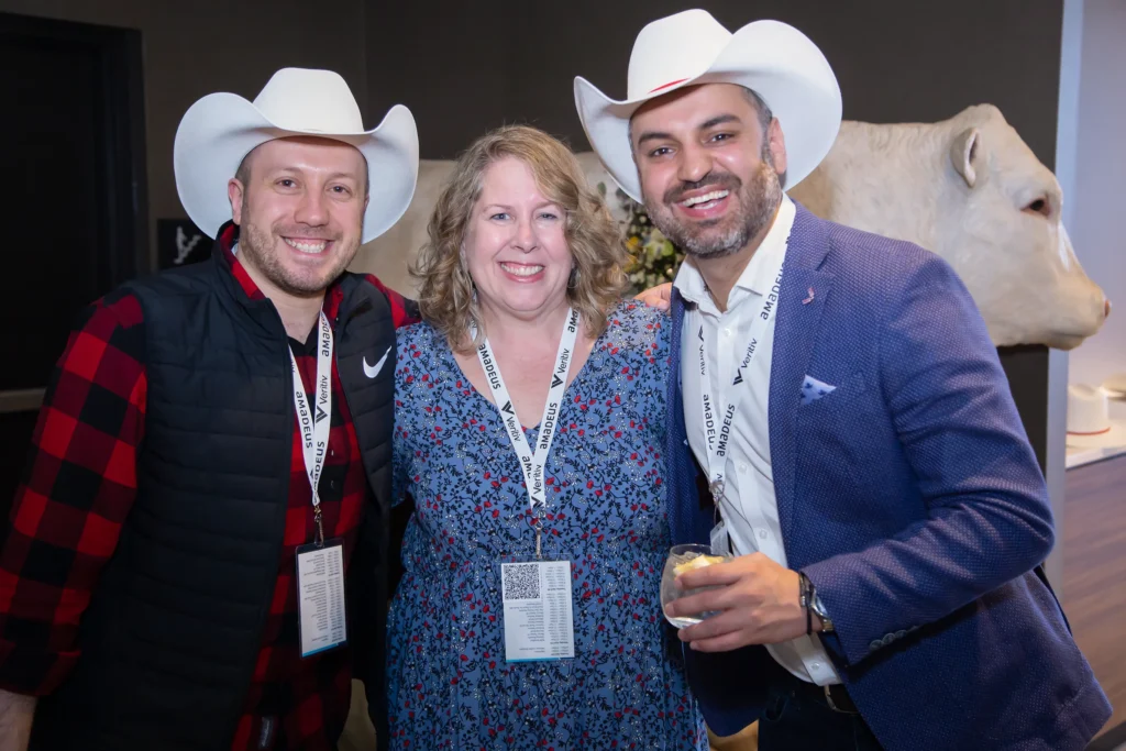 Three people wearing conference badges and cowboy hats smile together indoors; the two men stand on either side of a woman. One man wears a red plaid shirt and vest, the other a blue blazer, and a large white animal sculpture is in the background. - Jodi O Photography