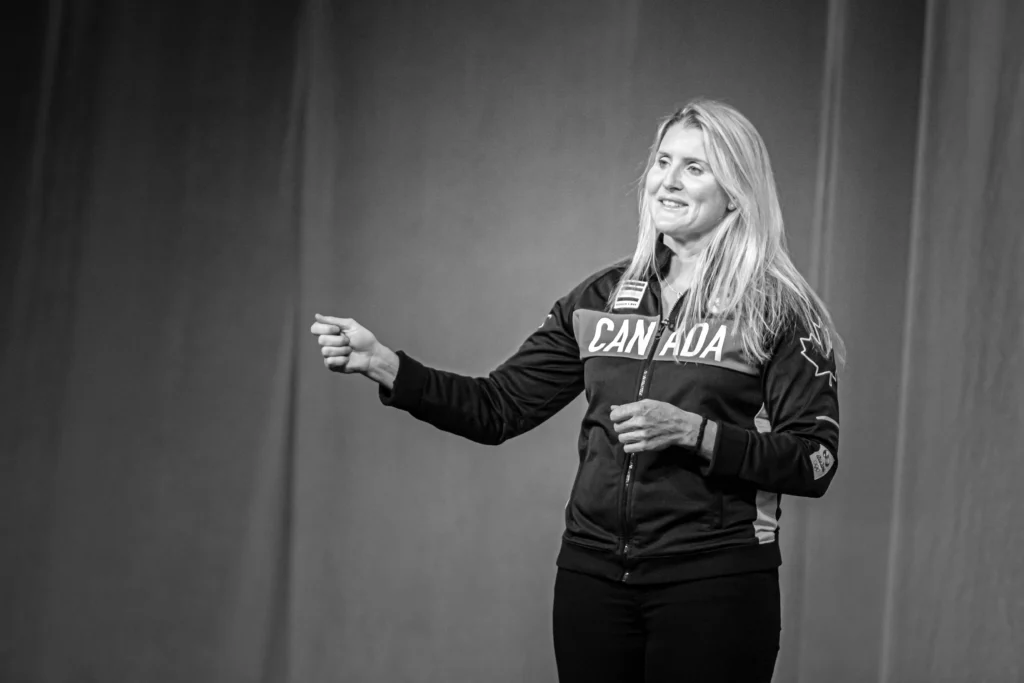 A woman wearing a "Canada" jacket stands on stage, smiling and gesturing with one hand. Captured in black and white by Calgary Photographers, the image radiates timeless charm. - Jodi O Photography
