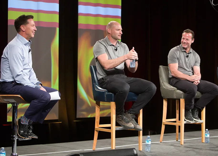 Three men sit on tall chairs on a stage during a panel discussion. One man holds a water bottle while speaking, and the others smile. Bottled water sits on the floor, and a colorful backdrop is behind them. - Jodi O Photography