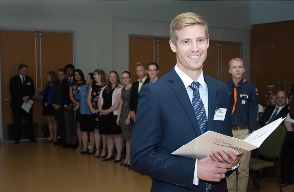 A man in a suit, holding a folder and smiling, stands in the foreground. Behind him, a group of people lines up against the wall, dressed formally, waiting in a large room with brown double doors. - Jodi O Photography