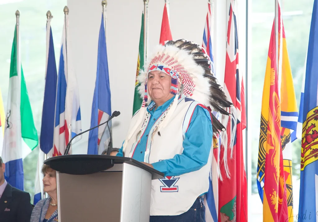 An Indigenous man wearing a feathered headdress and traditional attire stands at a podium, speaking. Various national and provincial flags are displayed in the background. - Jodi O Photography