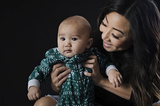 A woman with long dark hair smiles at a baby she is holding. The baby, in a green patterned outfit, looks slightly away from the camera. The dark background draws focus to their faces—perfect for Studio Family Photos Calgary. - Jodi O Photography