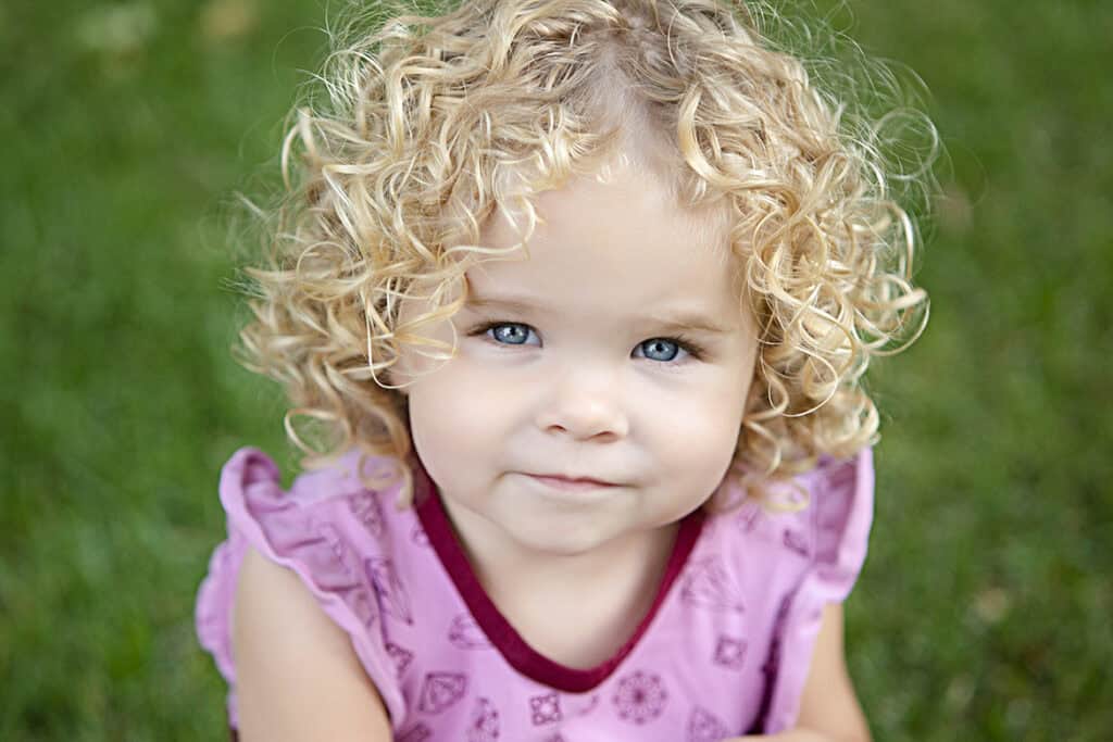 A young child with curly blonde hair and blue eyes, wearing a pink patterned shirt, looks up at the camera with a slight smile while sitting on green grass during an outdoor family photo session in Calgary. - Jodi O Photography
