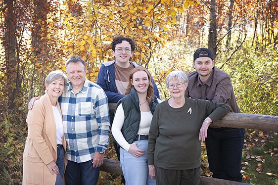 A group of six people, spanning three generations, stands outdoors in front of autumn trees. They are smiling, some leaning on a wooden fence, surrounded by colorful fall foliage—perfect for an Outdoor Family Photos Calgary session. - Jodi O Photography