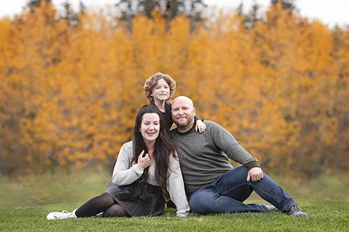 A smiling family of three poses on grass in front of vibrant autumn trees during their Outdoor Family Photos Calgary session. A boy stands behind a seated woman and man, all in casual fall clothing, surrounded by brilliant orange foliage. - Jodi O Photography