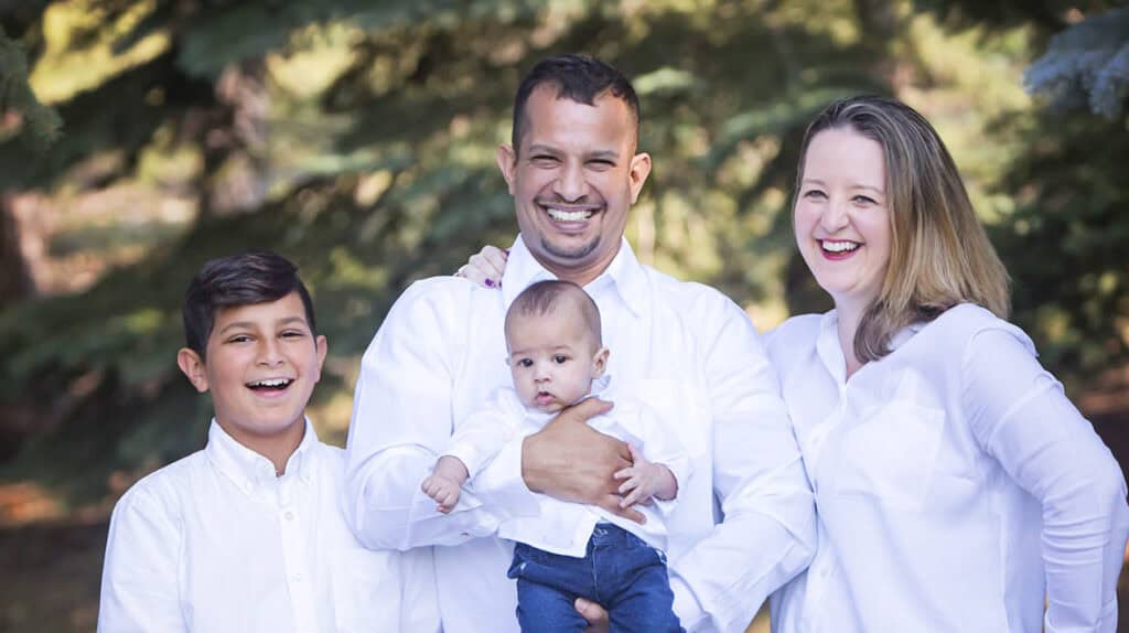 A family of four poses outdoors, smiling at the camera in front of green trees. All wear white shirts—perfect inspiration for Outdoor Family Photos Calgary, capturing natural moments with loved ones in a beautiful setting. - Jodi O Photography
