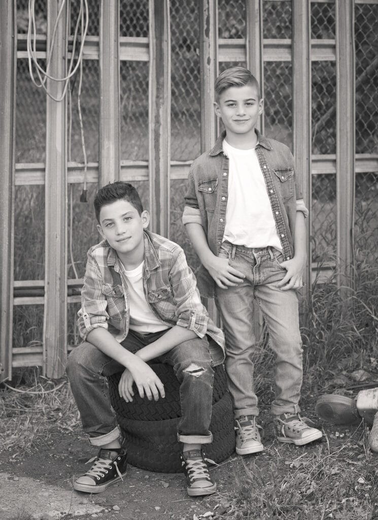 Two boys wearing jeans, t-shirts, and sneakers pose in front of a wire fence and stacked tires. Perfect for an outdoor family photos Calgary session, they look at the camera with relaxed expressions—one boy seated on the tires, the other standing beside him. - Jodi O Photography