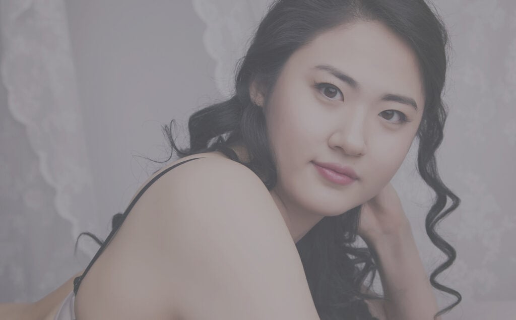 A woman with long, wavy black hair and smooth skin reclines and looks at the camera, resting her head on her hand. She wears a light-colored top, with lace curtains blurred in the background. - Jodi O Photography