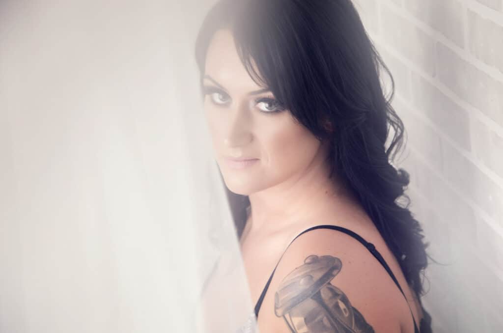 A woman with long dark hair and a visible tattoo on her shoulder looks at the camera. She stands beside a white brick wall, partly obscured by a sheer curtain, in soft, natural light. - Jodi O Photography