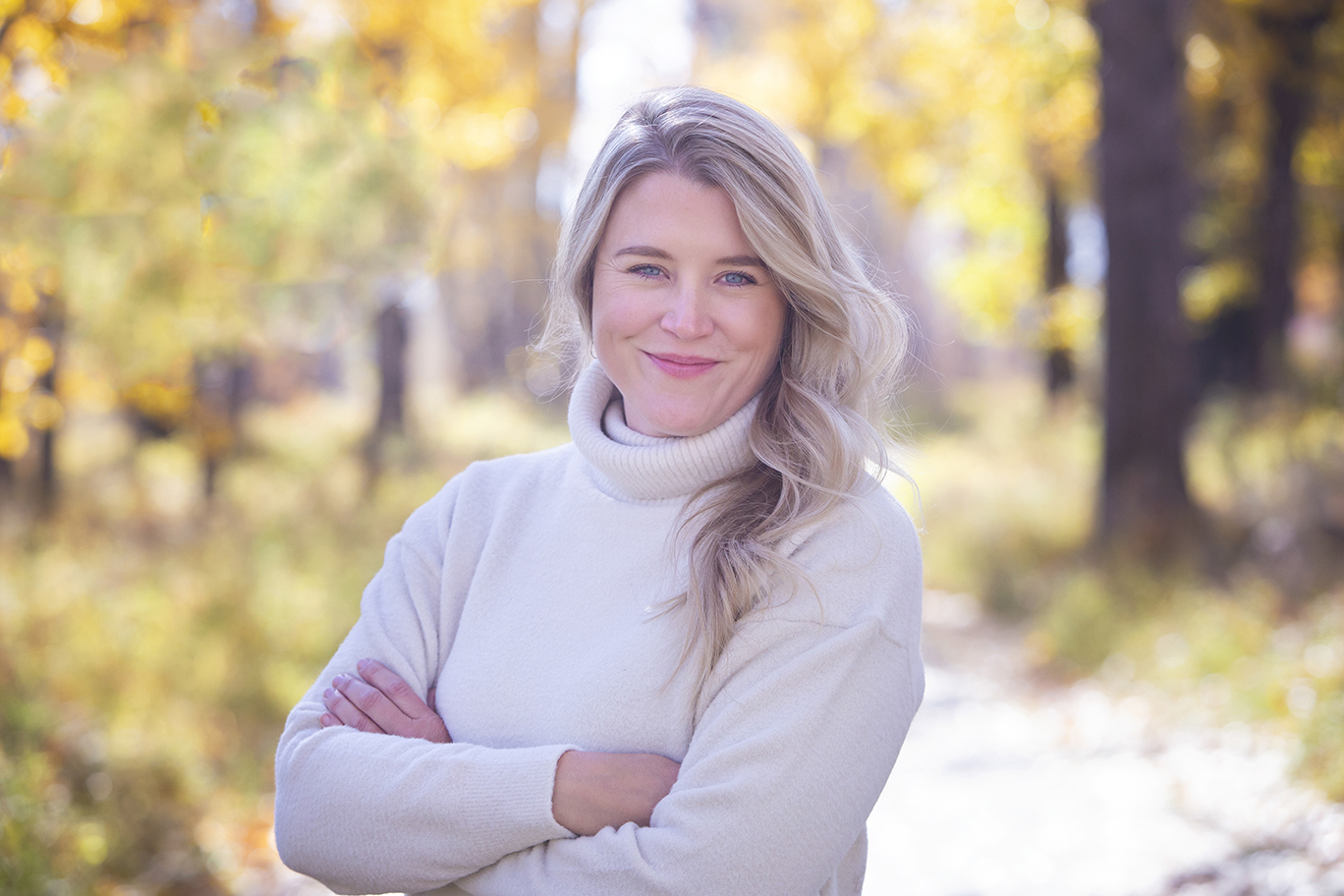A woman with long blonde hair, wearing a light turtleneck sweater, stands outdoors with her arms crossed, smiling, surrounded by trees with yellow autumn leaves. - Jodi O Photography