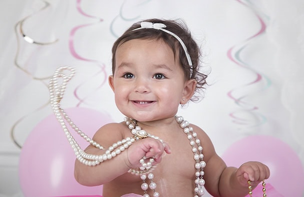 Smiling baby wearing a white headband and pearl necklaces, sitting in front of pink balloons and pastel streamers. Perfect for Studio Family Photos Calgary, the baby holds pearls and looks slightly to the side. - Jodi O Photography
