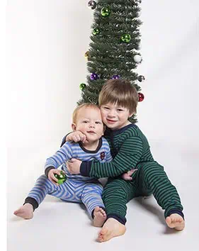 Two young children in striped pajamas sit and hug in front of a decorated Christmas tree. One child holds a green ornament, and both are smiling at the camera. - Jodi O Photography