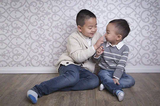 Two young boys sit on a wooden floor in front of a patterned wall. The older boy gently touches the younger boy’s nose, and both are smiling, wearing sweaters and jeans. - Jodi O Photography