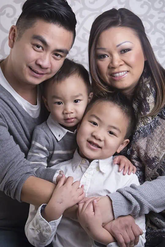 A smiling family of four poses closely together. Two adults stand behind, embracing two young boys in front who are also smiling. The background features a light, decorative pattern. - Jodi O Photography