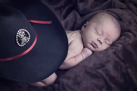 A newborn baby sleeps peacefully on a soft brown blanket next to a large black hat with a red band and a decorative badge. - Jodi O Photography
