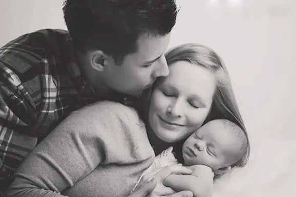 A man gently kisses a smiling woman on the forehead as she holds a sleeping newborn baby, all cuddled together in a warm, close embrace. The photo is in black and white. - Jodi O Photography