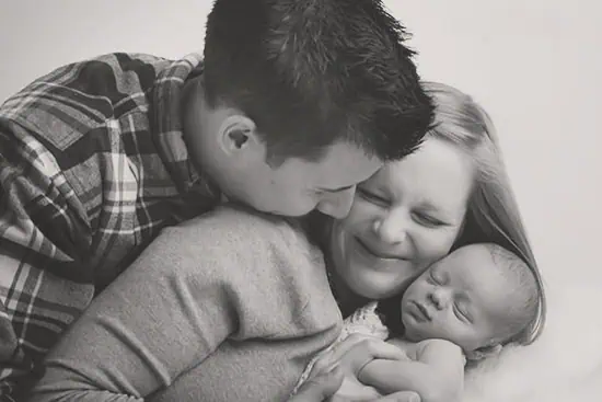A black-and-white photo of a happy couple cuddling closely with their newborn baby. The mother smiles with closed eyes while the father embraces both, and the baby sleeps peacefully in their arms. - Jodi O Photography
