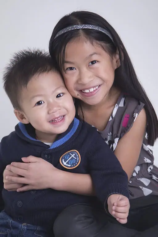 A young girl with long dark hair hugs a smiling toddler boy. Both children are sitting close together and looking at the camera against a plain background. The girl is wearing a headband and a gray dress; the boy wears a navy sweater. - Jodi O Photography