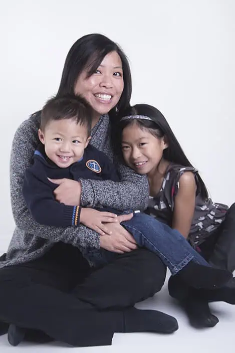 A woman sits on the floor, smiling and hugging a young boy and a young girl, both also smiling, in front of a plain white background. - Jodi O Photography