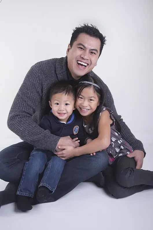 A man sits on the floor smiling and hugs a young boy and girl, who are also smiling. The group appears happy and close, photographed against a plain white background. - Jodi O Photography