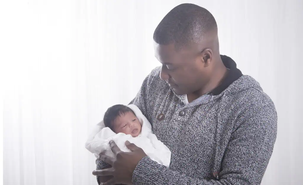 A man wearing a gray sweater gently holds a newborn baby wrapped in a white blanket, looking at the baby with a soft expression, against a light, softly blurred background. - Jodi O Photography