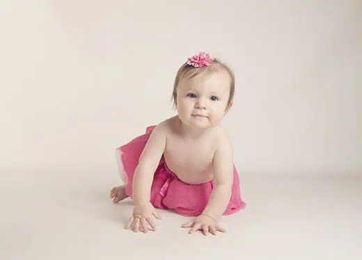 A baby wearing a pink tutu skirt and a pink flower headband crawls on a light-colored floor with a neutral background, looking toward the camera. - Jodi O Photography