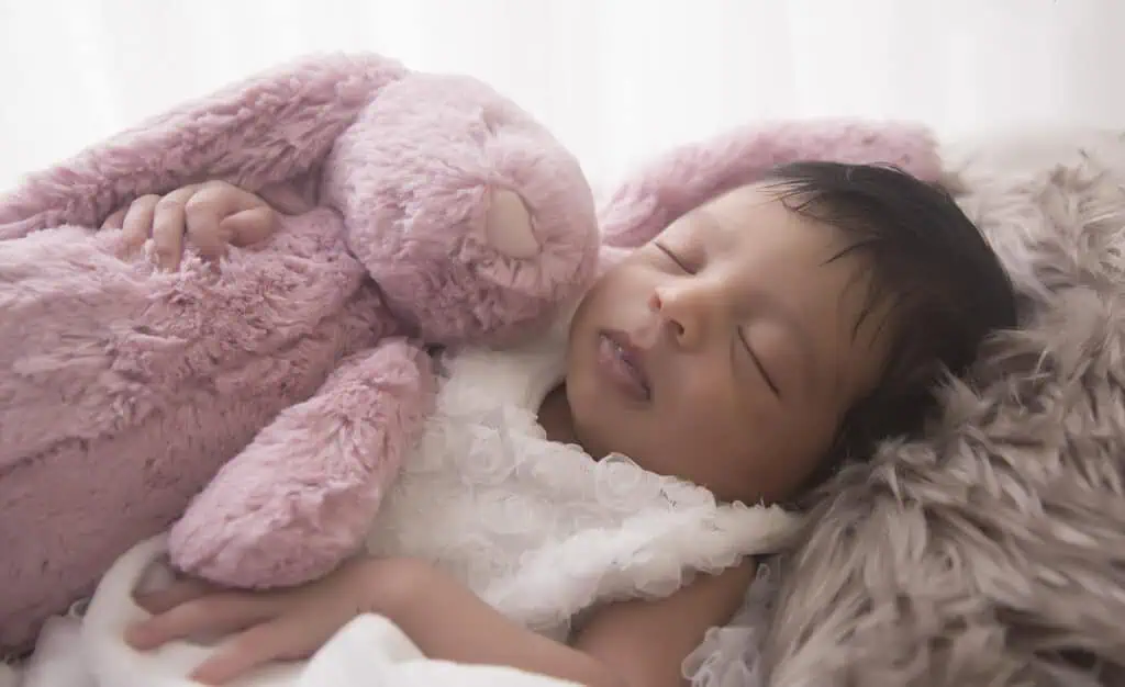 A newborn baby sleeps peacefully while hugging a soft pink stuffed bunny, nestled in cozy blankets with a gentle, warm light in the background. - Jodi O Photography