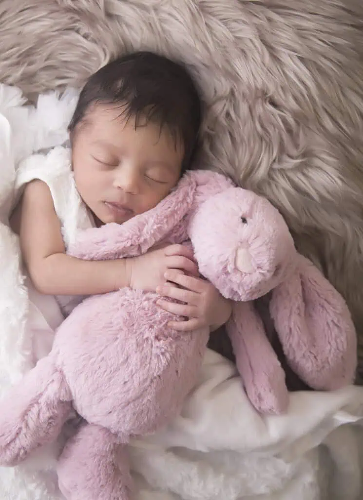 A sleeping newborn baby cuddles a soft pink stuffed bunny, lying on a fluffy beige blanket with a white blanket partially covering them. The baby looks peaceful and content. - Jodi O Photography