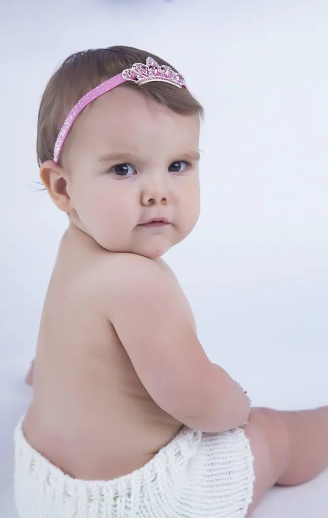 A baby wearing a white knitted diaper cover and a pink headband with a tiara sits against a light background, looking over their shoulder towards the camera. - Jodi O Photography