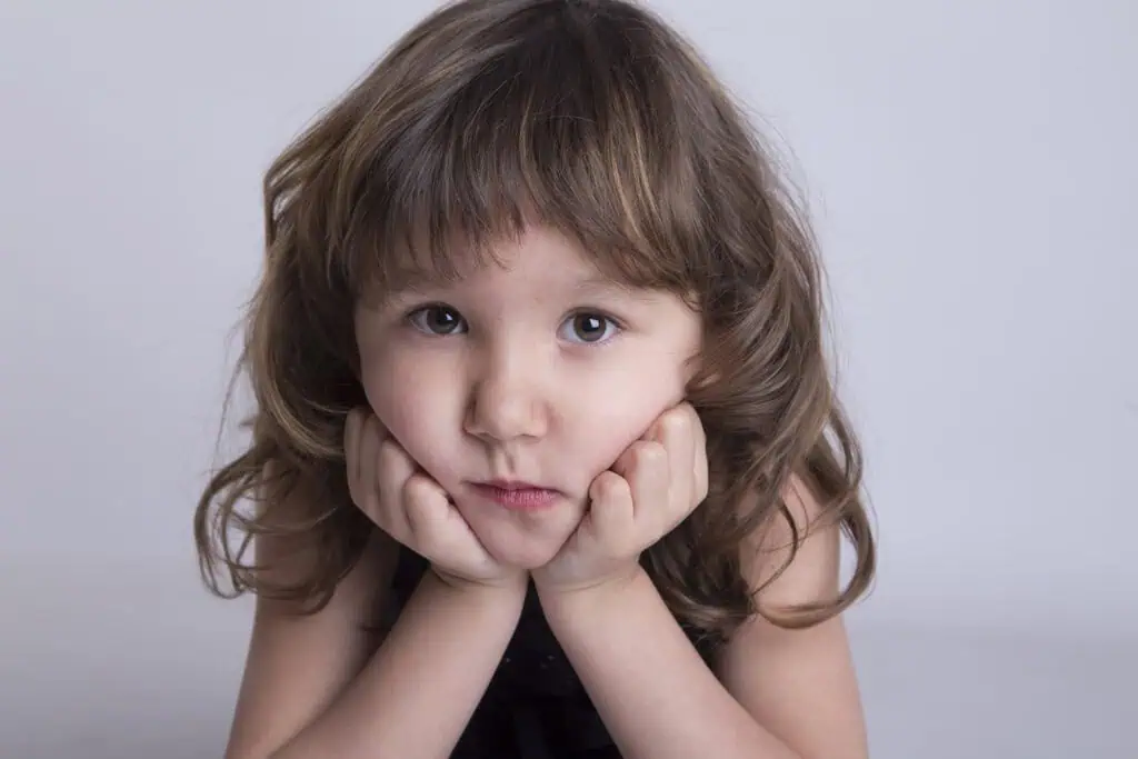 A young child with wavy brown hair rests their chin on their hands, looking directly at the camera with a serious expression against a plain light background. - Jodi O Photography