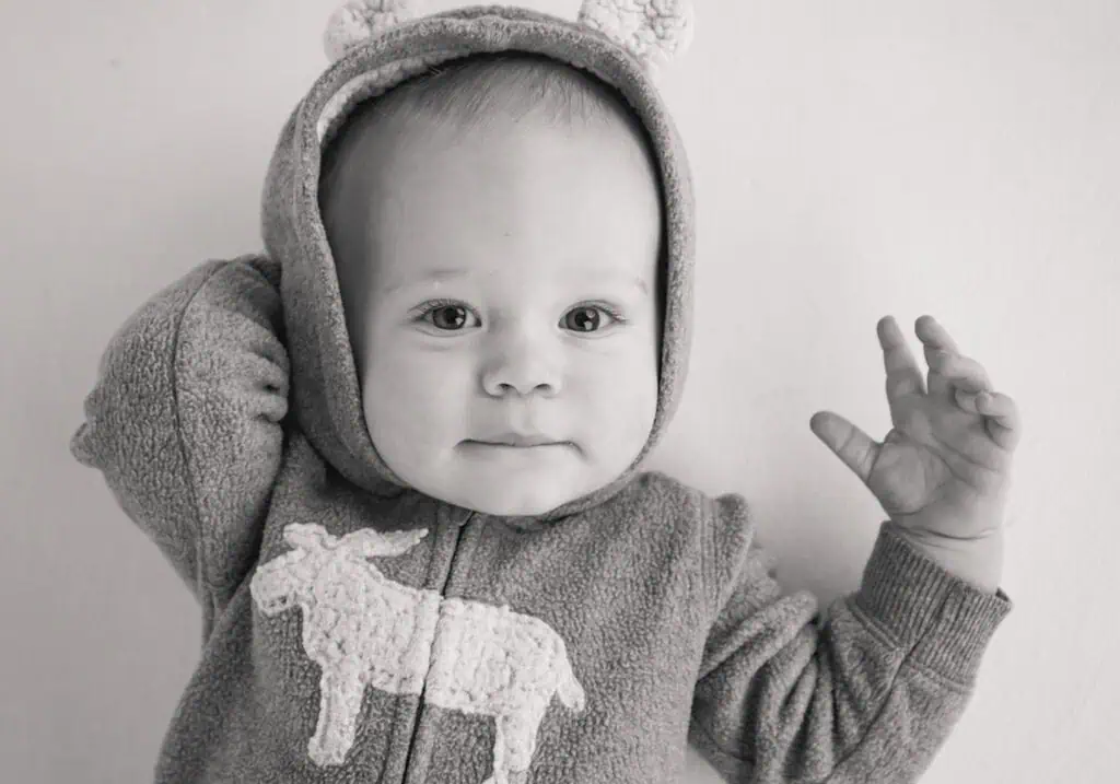Black and white photo of a baby wearing a hooded onesie with animal ears and a donkey design on the front, looking at the camera with one arm raised and a slight smile. - Jodi O Photography