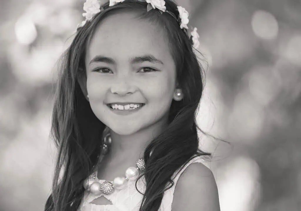 A young girl with long hair smiles at the camera, wearing a floral headband, pearl necklace, and a sleeveless dress. The background is softly blurred, creating a gentle, dreamy effect. The image is in black and white. - Jodi O Photography