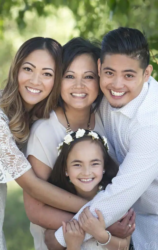 Four people, including a young girl wearing a flower crown, two women, and a man, stand close together outdoors, smiling brightly as they embrace each other in a warm group hug. Greenery is blurred in the background. - Jodi O Photography