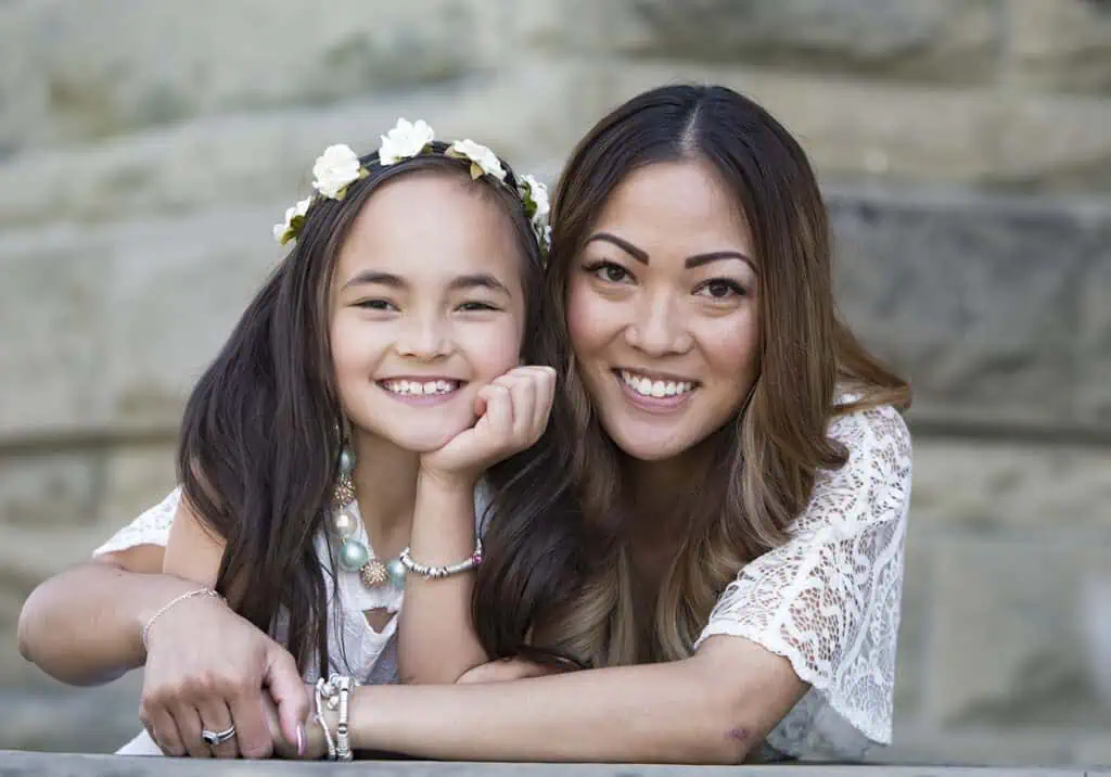 A smiling woman and young girl with long dark hair lean on a stone surface outdoors. Both wear white tops; the girl has a flower crown and turquoise necklace. They appear happy and close, posing together. - Jodi O Photography