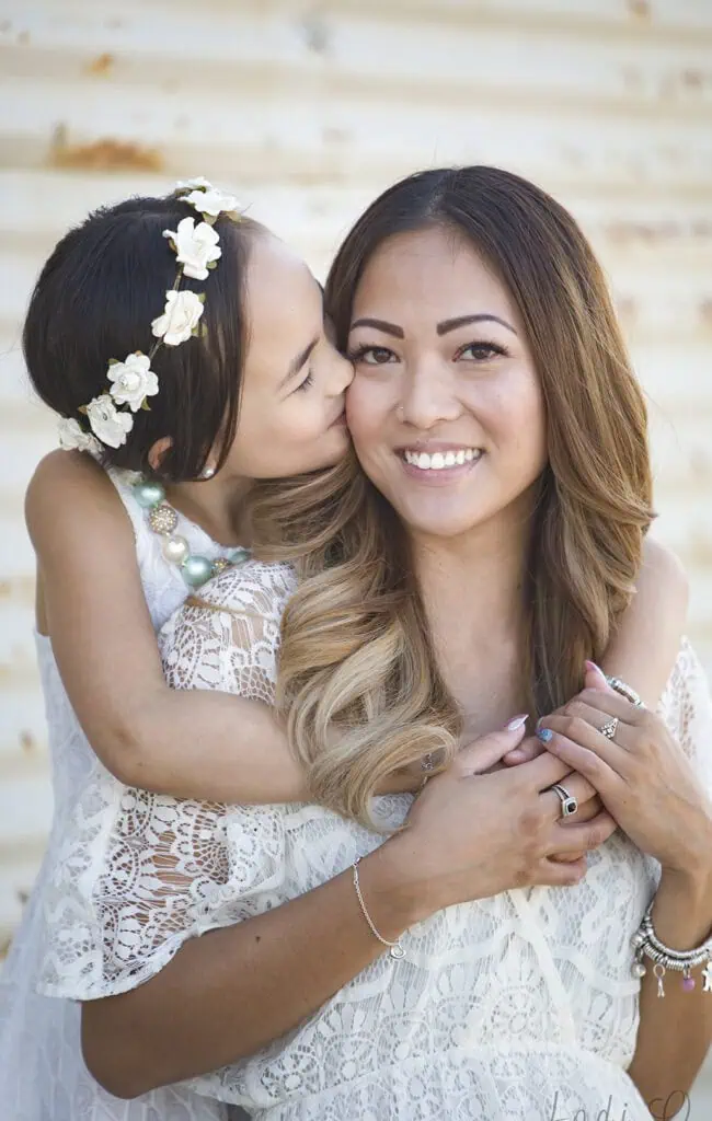 A young girl wearing a white flower headband kisses a smiling woman on the cheek from behind. Both are dressed in white lace outfits, and the background is a light, rustic wall. - Jodi O Photography