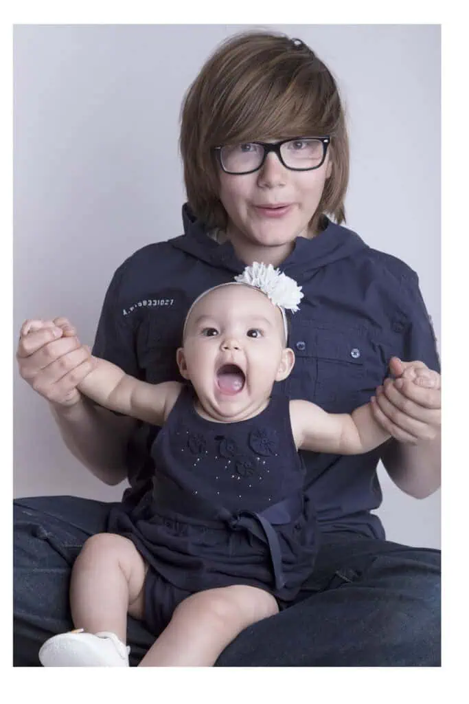 A teenage boy with glasses sits behind a smiling baby girl wearing a flower headband and a navy outfit. He holds her hands as she opens her mouth wide, showing excitement. Both appear happy, posing together against a plain backdrop. - Jodi O Photography