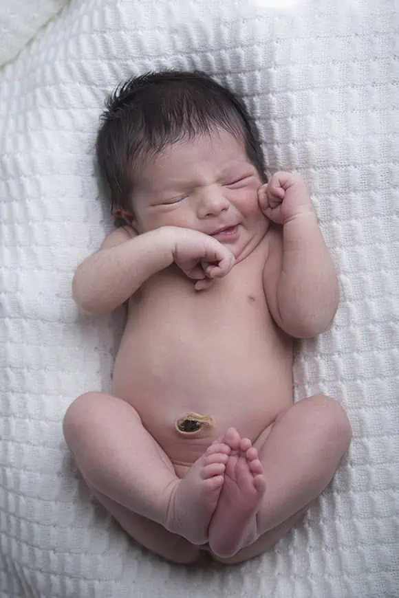 A newborn baby lies on a soft white textured blanket with eyes closed, fists near their face, and legs bent. The baby’s umbilical cord stump is still visible. - Jodi O Photography