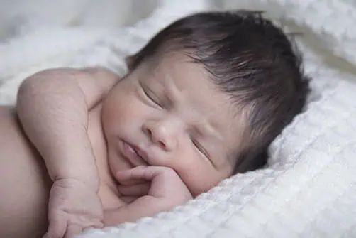 A newborn baby with dark hair sleeps peacefully on a soft white blanket, resting one hand against their cheek. - Jodi O Photography
