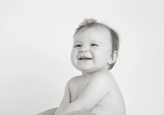 Black and white photo of a smiling baby sitting with bare shoulders, looking slightly to the side. The baby has light hair with a small flower accessory and a joyful expression. The background is plain and light-colored. - Jodi O Photography