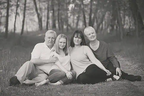 Four people, two older adults and two younger women, sit closely together on the ground in a wooded outdoor setting, smiling at the camera. The image is in black and white. - Jodi O Photography