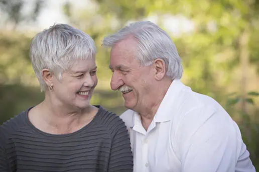 An elderly couple sits outdoors, smiling and looking at each other joyfully. The woman has short gray hair and wears a gray top; the man has white hair and a mustache, and wears a white shirt. Greenery is blurred in the background. - Jodi O Photography