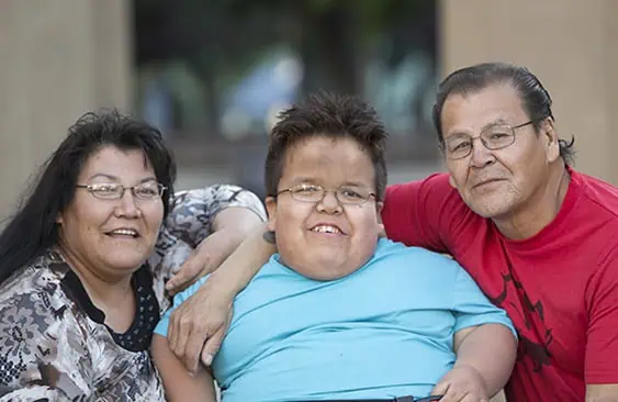 Three people sit close together outdoors, smiling at the camera. The person in the center wears a turquoise shirt and glasses, with an adult on each side, one in a patterned blouse and the other in a red shirt. - Jodi O Photography