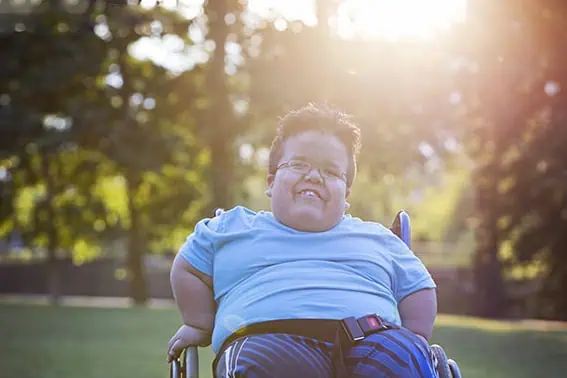 A smiling person in a blue shirt sits in a wheelchair outdoors on a sunny day, with green trees and bright sunlight in the background. - Jodi O Photography