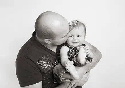A man lovingly holds and kisses a smiling baby on the cheek. The baby has a flower headband and is wearing a patterned dress and jeans. The photo is in black and white with a plain background. - Jodi O Photography