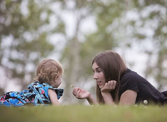 A woman and a young child lie on the grass facing each other, sharing a sweet moment outdoors with trees blurred in the background—perfect inspiration for your next outdoor family photos Calgary session. - Jodi O Photography