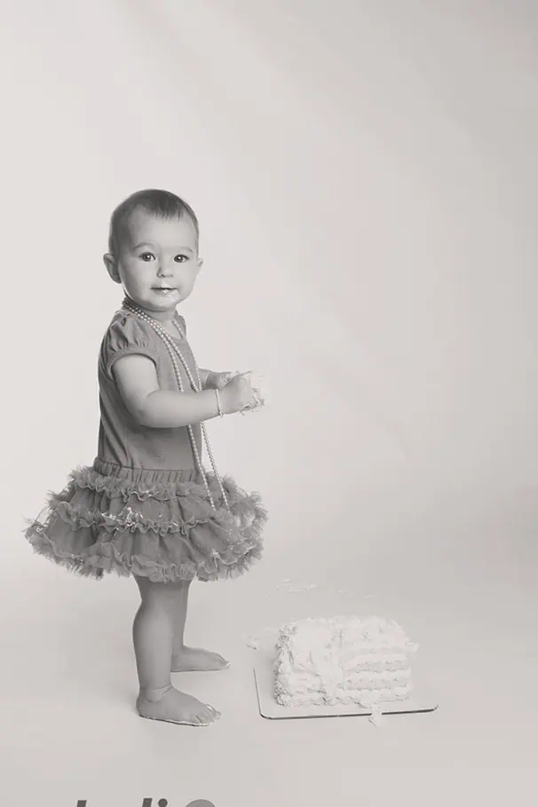 A smiling toddler wearing a frilly skirt and necklace stands barefoot beside a partially eaten cake on the floor. The background is plain and light-colored. The photo is in black and white. - Jodi O Photography