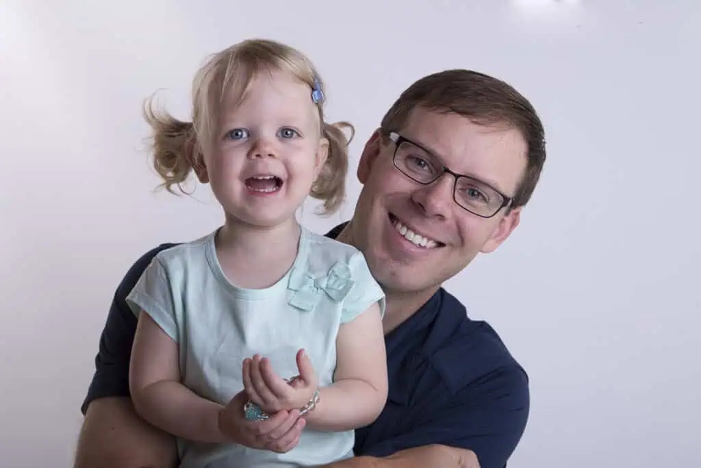 A smiling man wearing glasses hugs a cheerful young girl with blonde pigtails and a blue hair clip. Both look directly at the camera against a plain white background. - Jodi O Photography