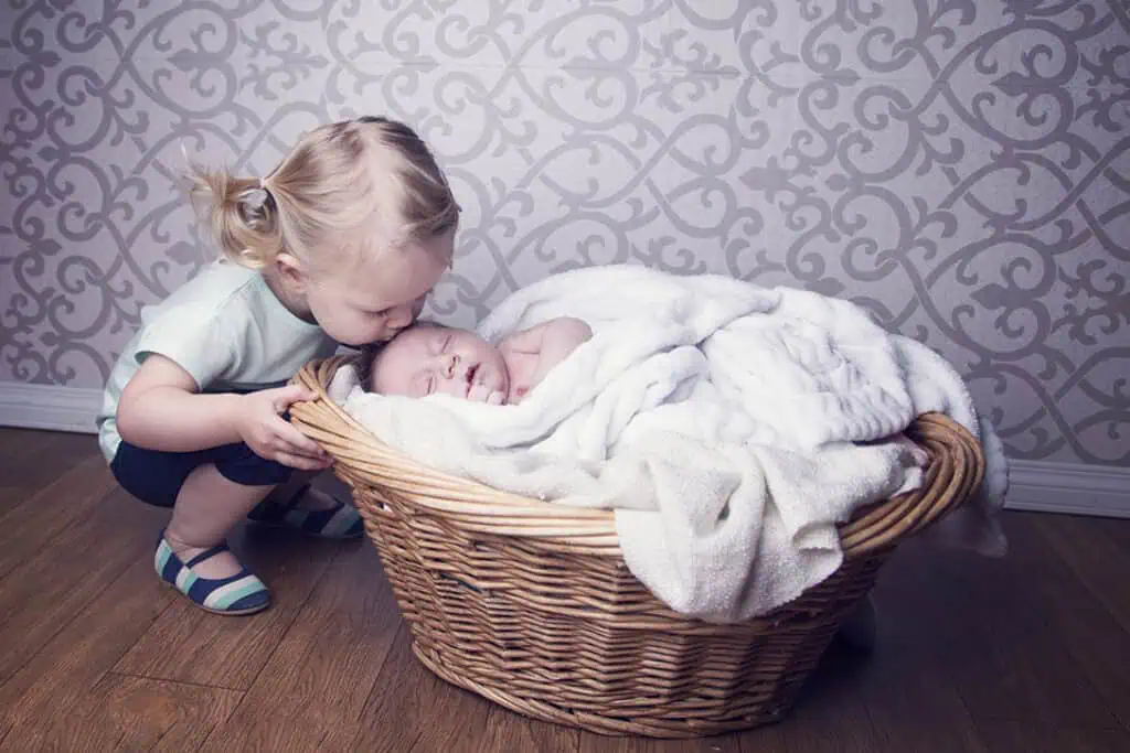 A young child gently kisses the forehead of a sleeping baby who is wrapped in a white blanket and lying in a wicker basket on a wooden floor, with a patterned wallpaper in the background. - Jodi O Photography